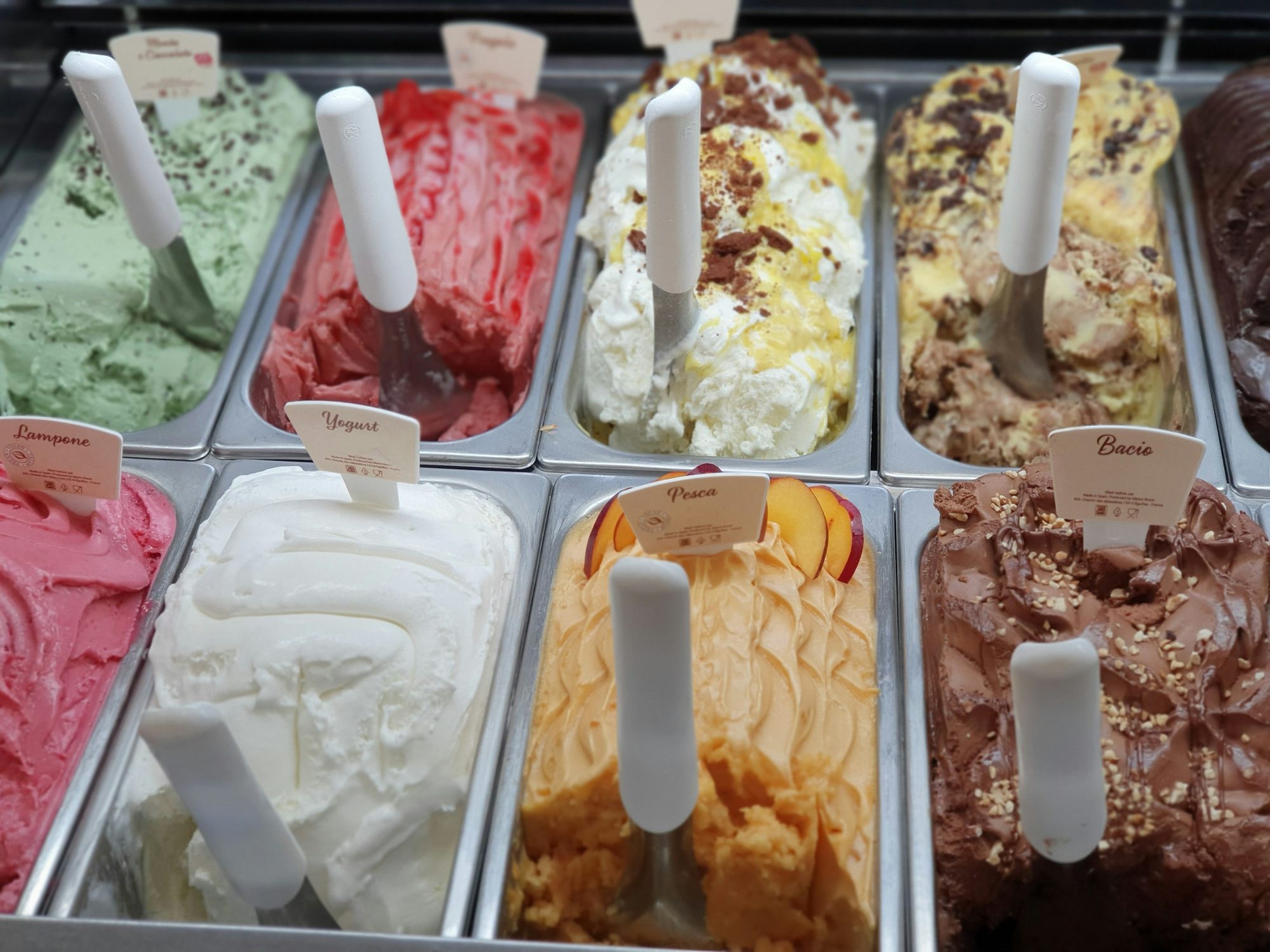 Une rangée de bacs de gelato colorés — pistache, fraise, chocolat, vanille — avec leurs spatules